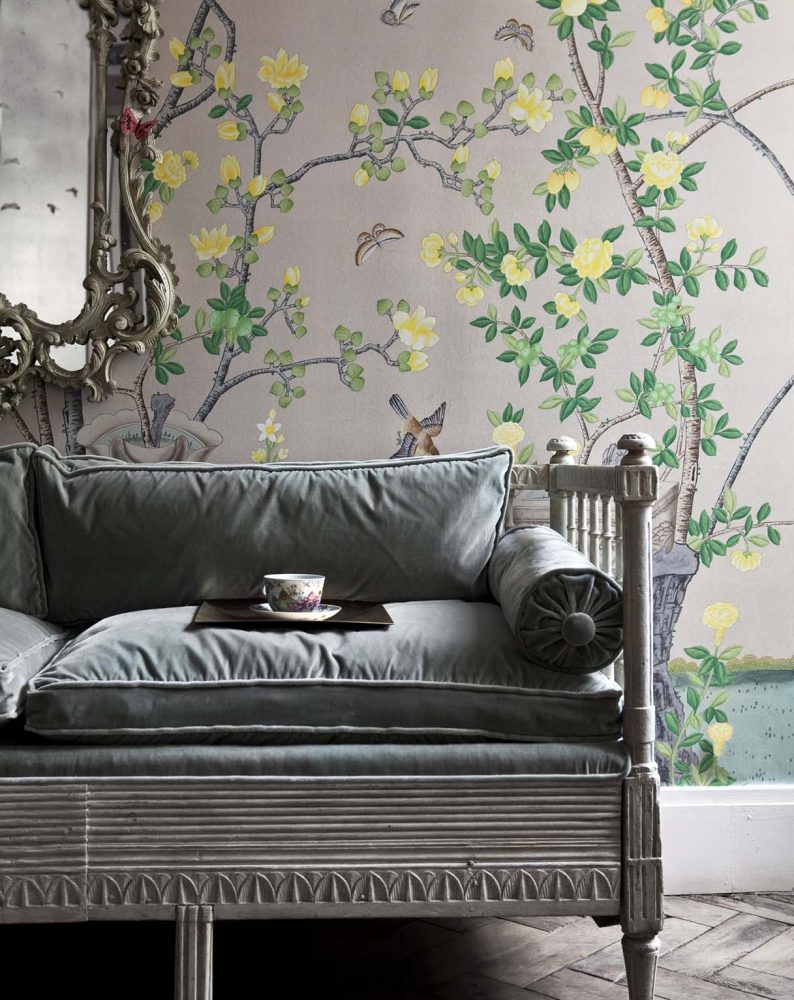 A tea cup and saucer sit on a pale gray velvet sofa, with de Gournay’s ‘Jardinieres Citrus Trees’ on metallic Chinese rice paper on the walls behind it
