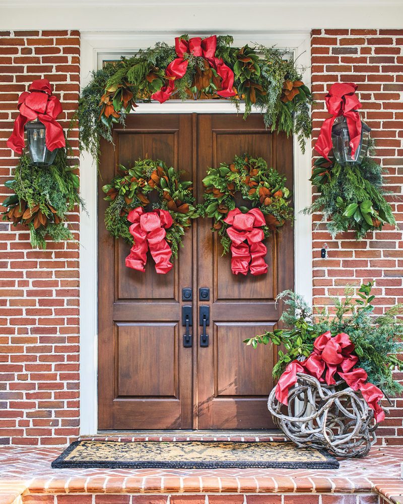 A grand set of natural wood doors on a brick on, with a wreath on each door, bundles of greenery on lantern, an arch of greenery over the doorway. To the side of the door, Canaan Marshall also adorned a grapevine sculpture with greenery. Large red bows finish each decorative element.
