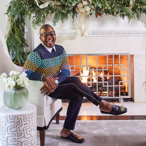 Atlanta floral designer Canaan Marshall, wearing a stylish crochet sweater, tailored dark pants a thin light strip along the outer seam, and loafers. The mantel is elaborately decorated with greenery and gold ribbon for the holidays. 