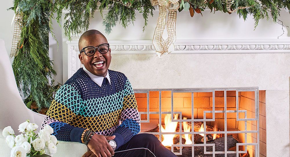 Atlanta floral designer Canaan Marshall, wearing a stylish crochet sweater, tailored dark pants a thin light strip along the outer seam, and loafers. The mantel is elaborately decorated with greenery and gold ribbon for the holidays.
