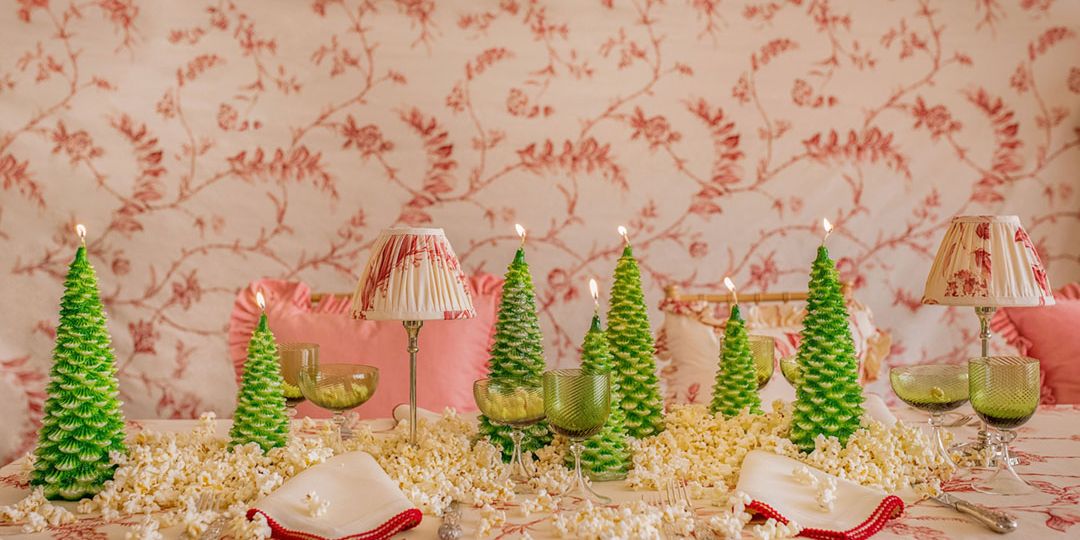 Christmas tree-shaped candles adorn a table dressed in a soft red and white botanical tablecloth print, napkins, and small lamp. Popcorn decorates the table, too.