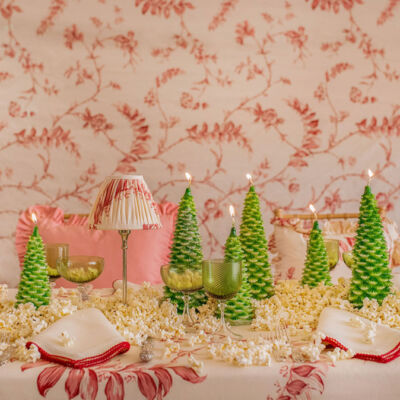 Christmas tree-shaped candles adorn a table dressed in a soft red and white botanical tablecloth print, napkins, and small lamp. Popcorn decorates the table, too.