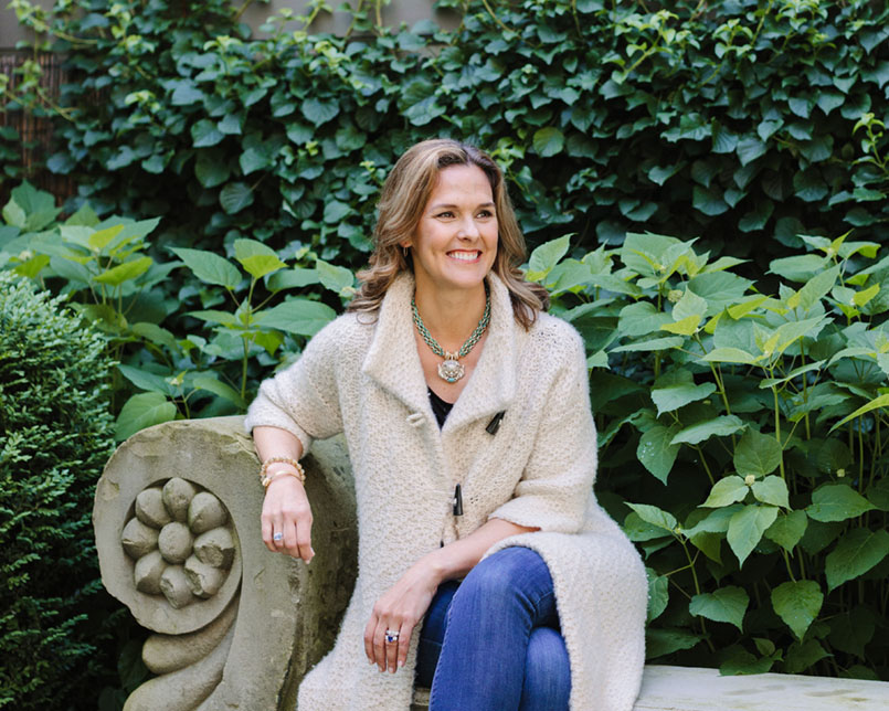 Jewelry designer Clara Williams, wearing a long neutral-colored cardigan sweater and jeans, sits on an elegant concrete bench against a lush green background