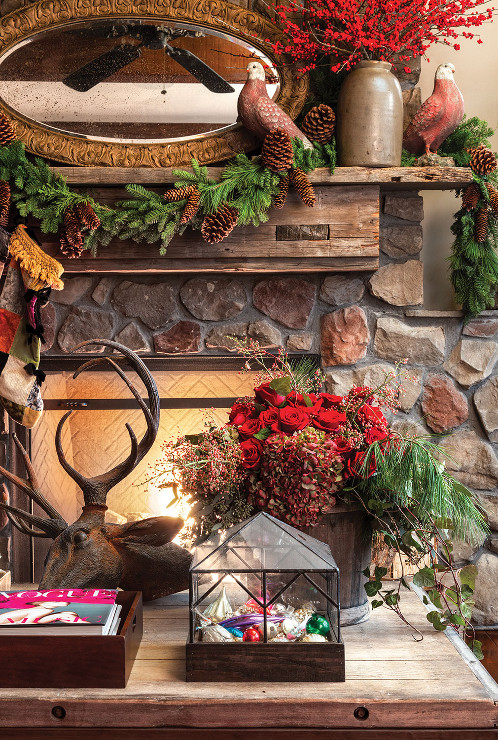 The scene includes an iron sculpture of a deer head with antlers, a leaded glass box holding vintage ornaments, and an red rose and evergreen floral arrangement on the coffee table. On the stone fireplace behind it, the rustic wood mantel is decorated with a mixed evergreen garland, two decorative partridges, and pottery vessel field with branches of red berries
