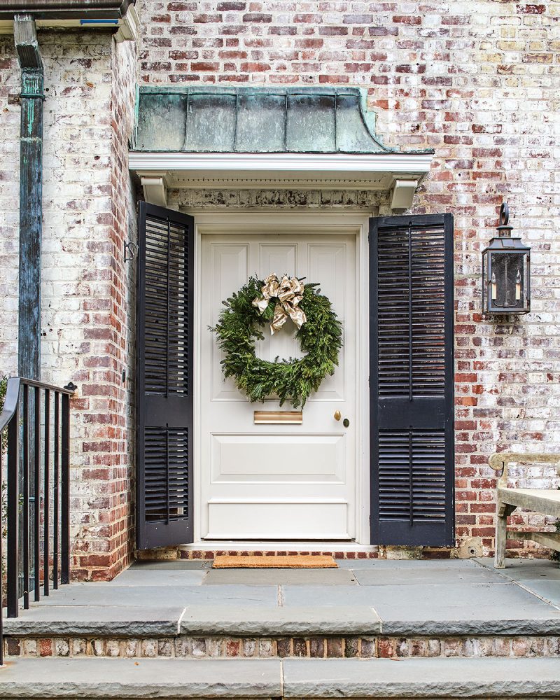 A wreath hangs on the white wooden door, which is flanked by black shutters. Above is an aged copper overhang. The photo also gives a closer look at the limewashed brick exterior of Jane Schwab’s Colonial-style home.