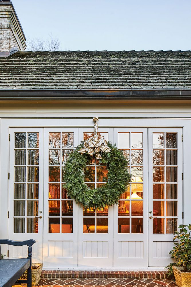 A large green holiday wreath with gold bow hangs on a section a mullioned windows between the pair of narrow French doors.