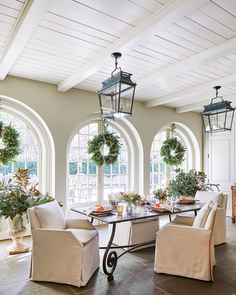 The light and airy garden room features a painted wood ceiling with exposed beams, three sets of arched French doors, and a natural wood dining table with a scrolled metal base, surrounded by natural linen slip-covered chairs. Two large black lanterns provide lighting.