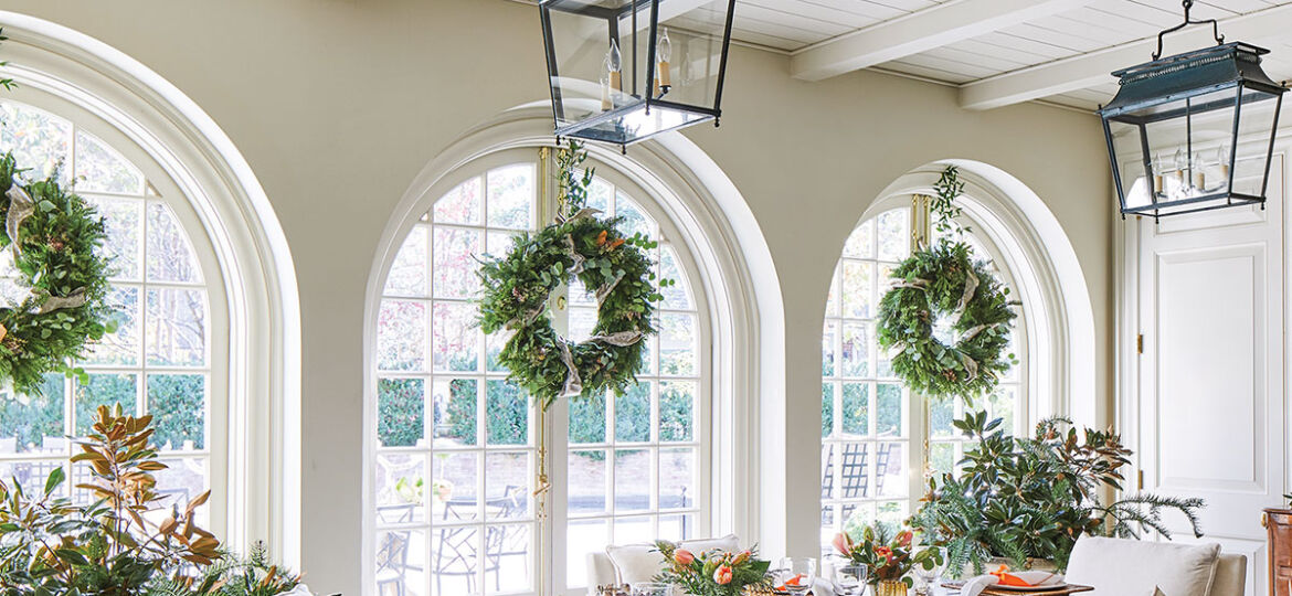 The light and airy garden room features a painted wood ceiling with exposed beams, three sets of arched French doors, and a natural wood dining table with a scrolled metal base, surrounded by natural linen slip-covered chairs. Two large black lanterns provide lighting.