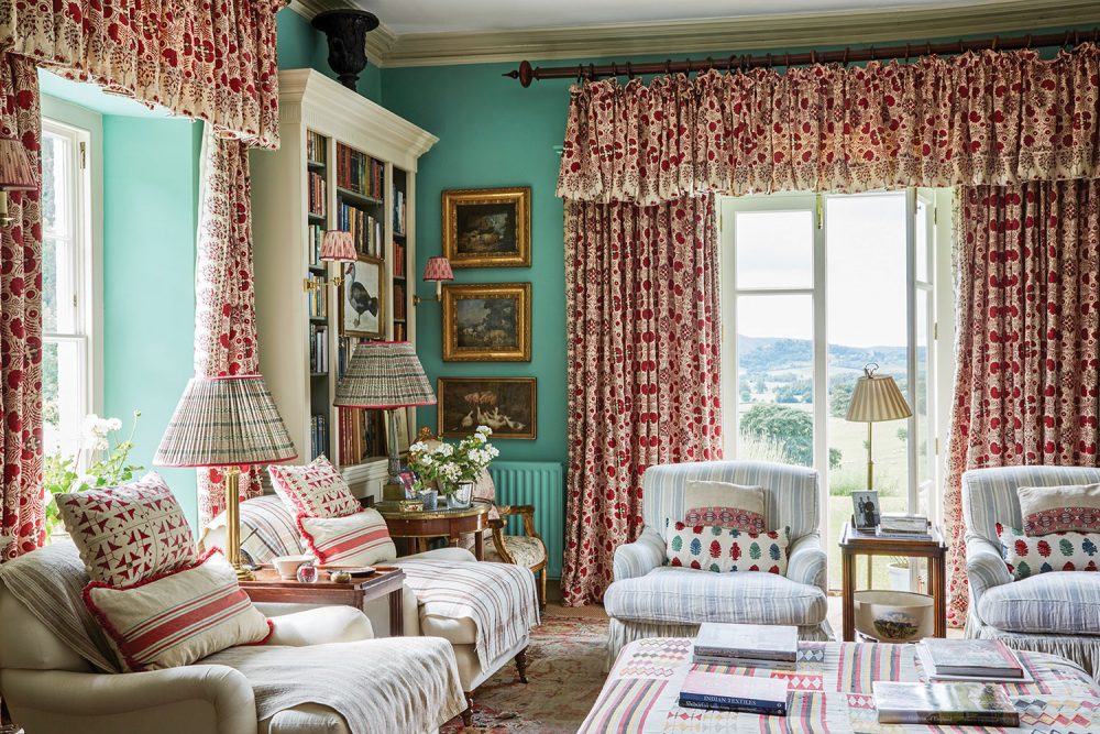 The aqua wall color contrasts with the muted reds featured prominently in various prints on the draperies and throw pillows. Comfortable reading chairs wear soft white upholstery and slipcovers with a pale stripe pattern