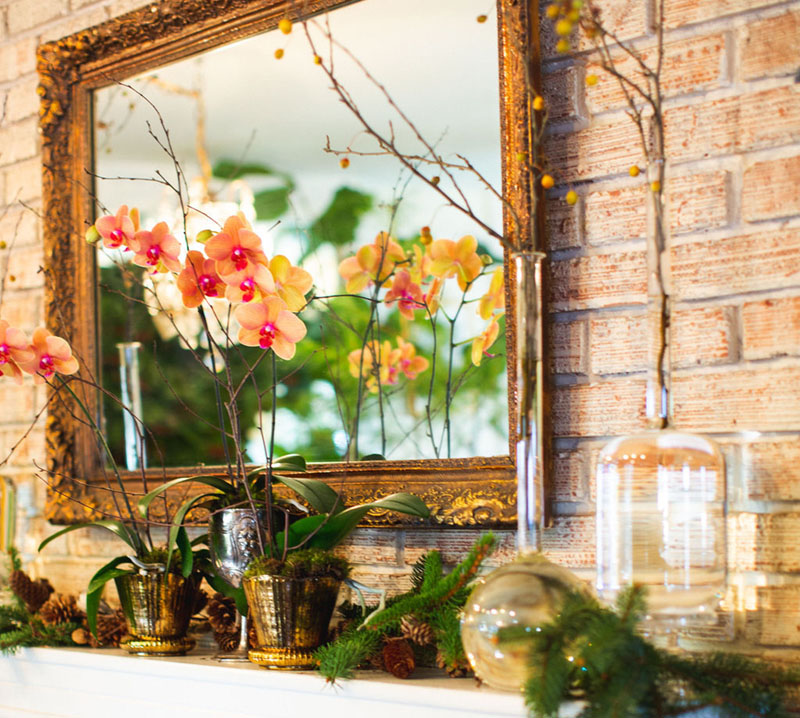Potted orchids, branches of berries displayed in vintage glass bottles, and evergreen clippings on a mantel beneath a gilt antique mirror
