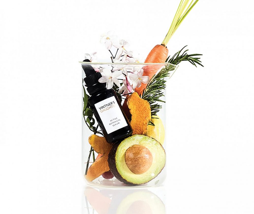 Product shot showing a serum in small dark glass bottle with Vintner’s Daughter logo, placed in a glass beaker with some of its ingredients - avocado, carrot, flowers, and herbs.