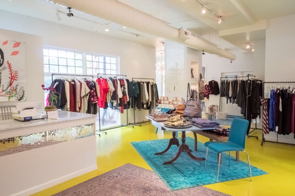 Scout Boutique's light and bright interior with painted yellow floors, turquoise and neutral oriental rungs, an antique pedestal dining table holding folded cloths, and long free-standing racks of clothes around the perimeter 
