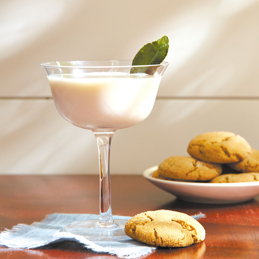 A creamy white gin-based cocktail garnished with a lime leaf next to a plate of ginger snap cookies