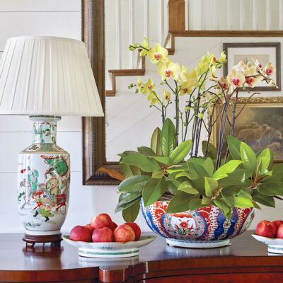 Lamp, phaleanopsis orchid arrangement and bowl of fruit on sideboard.