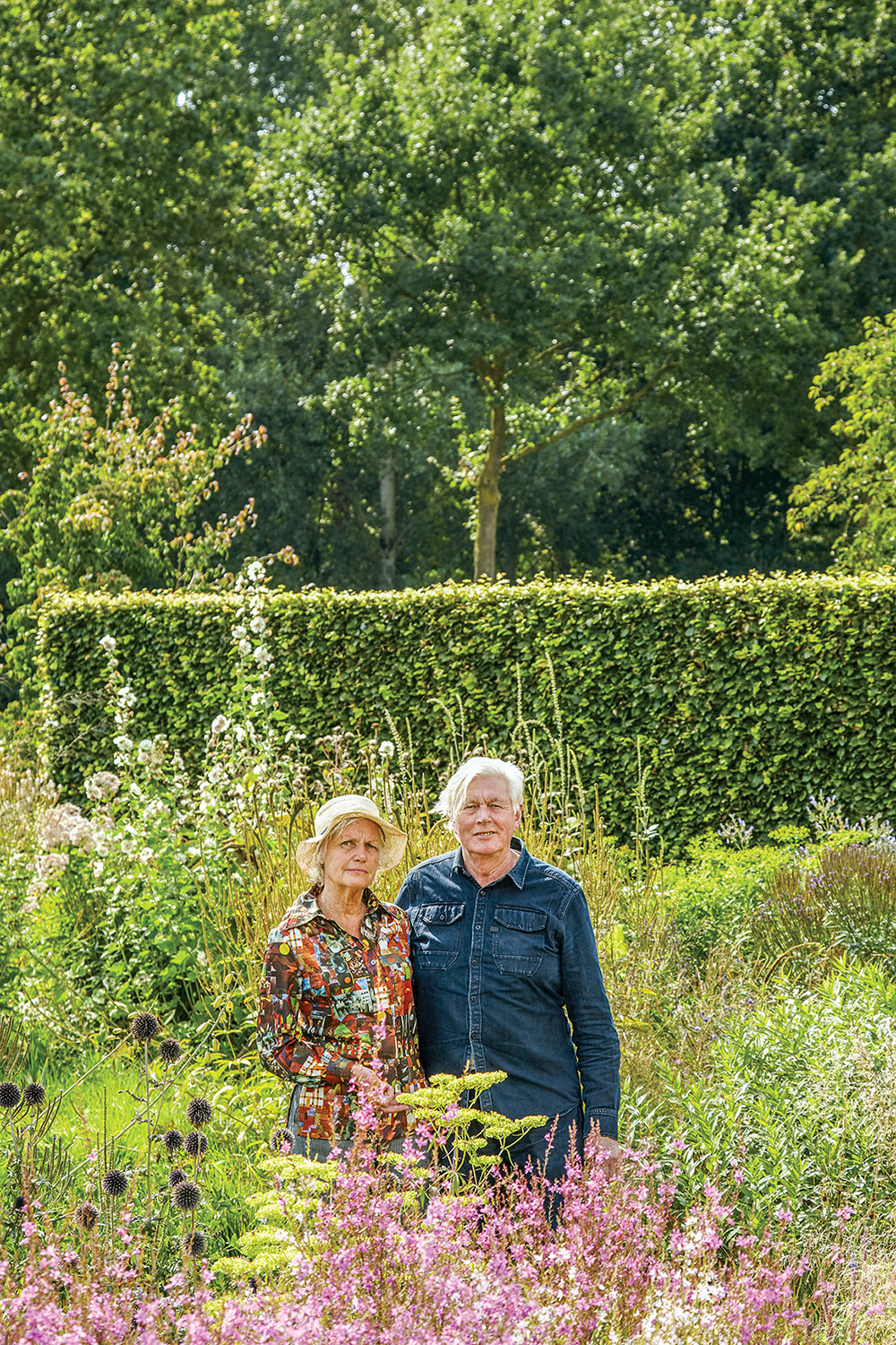 Portrait of the Dutch landscape designer (left), with silver hair and wearing a long sleeve denim buttonup, alongside his wife, wearing a colorful top and tan brimmed hat, in their private garden surrounded by grasses and perennial flowers