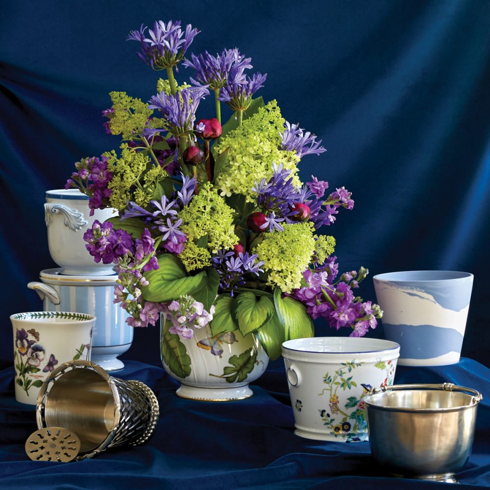A collection of cachepot, with a vibrant purple and green floral arrangement filling one of the vessels in the center, against a dark blue velvet backdrop