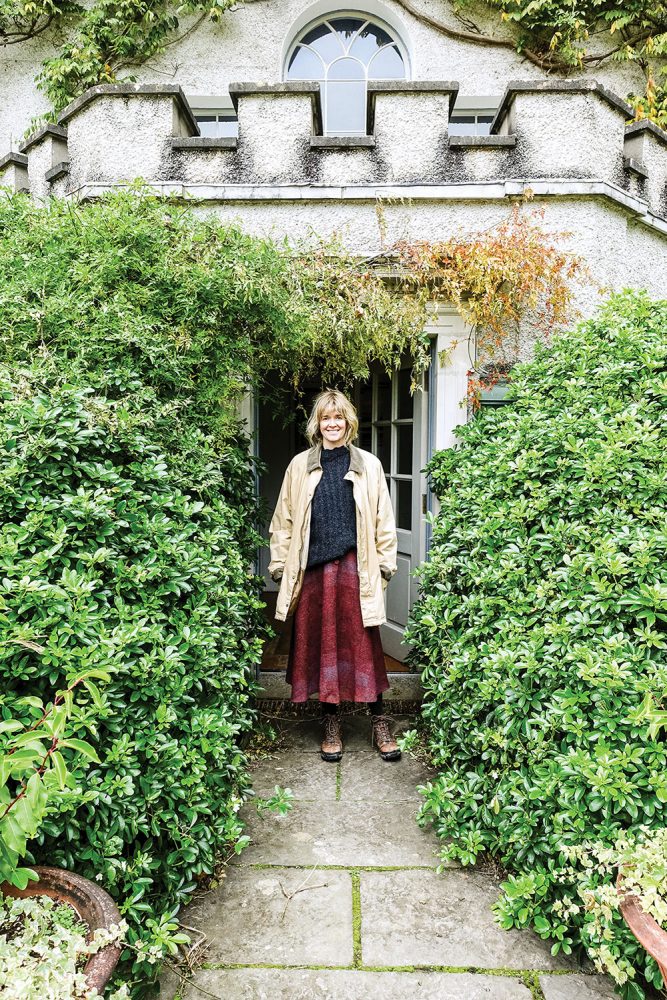 Glin Castle heiress Catherine FitzGerald, with chin-length wavy blond hair and a pleasant smile, wears rugged boots, a light tan farm jacket, a dark cable-knit sweater; and a full, ankle-length, burgundy skirt in a paisley print