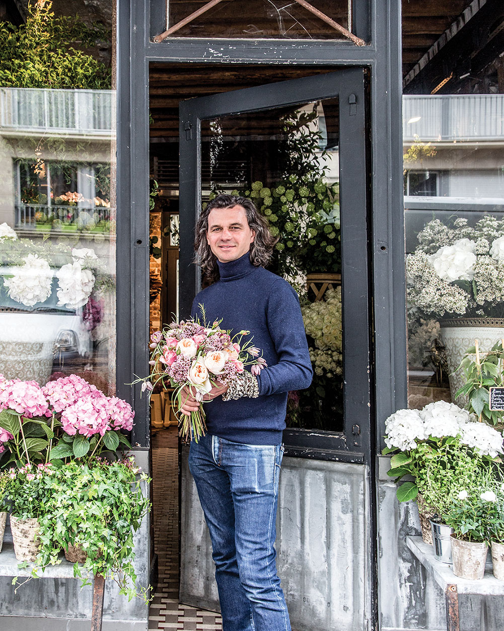 Parisian florist Eric Chauvin, wearing in worn jeans and navy turtleneck, stands at the open door of his French flower shop, holding a bouquet. On either side, window boxes are planted with pink and white flowering hydrangeas and trailing ivy.