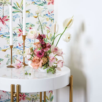 An ikebana arrangement and a pair of simple brass candlesticks with white tapers sit on a modern, white, circular occasional table with brass legs. A panel of floral fabric hangs in the background