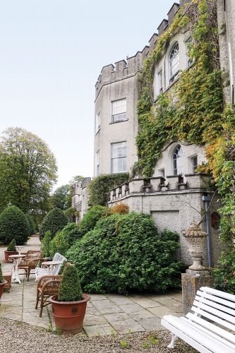 Vacationing in Ireland: View of Glin Castle's crenellated exterior, partially covered in climbing vines and bordered by green shrubs. At the base of the shrubs, ample benches for houseguests face out toward the garden view.