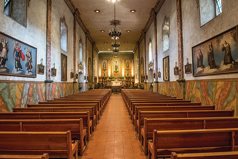 View of Old Santa Barbara Mission sanctuary, with red tile floors, natural wood pews, and art hung all long the stucco walls beneath high, deep windows