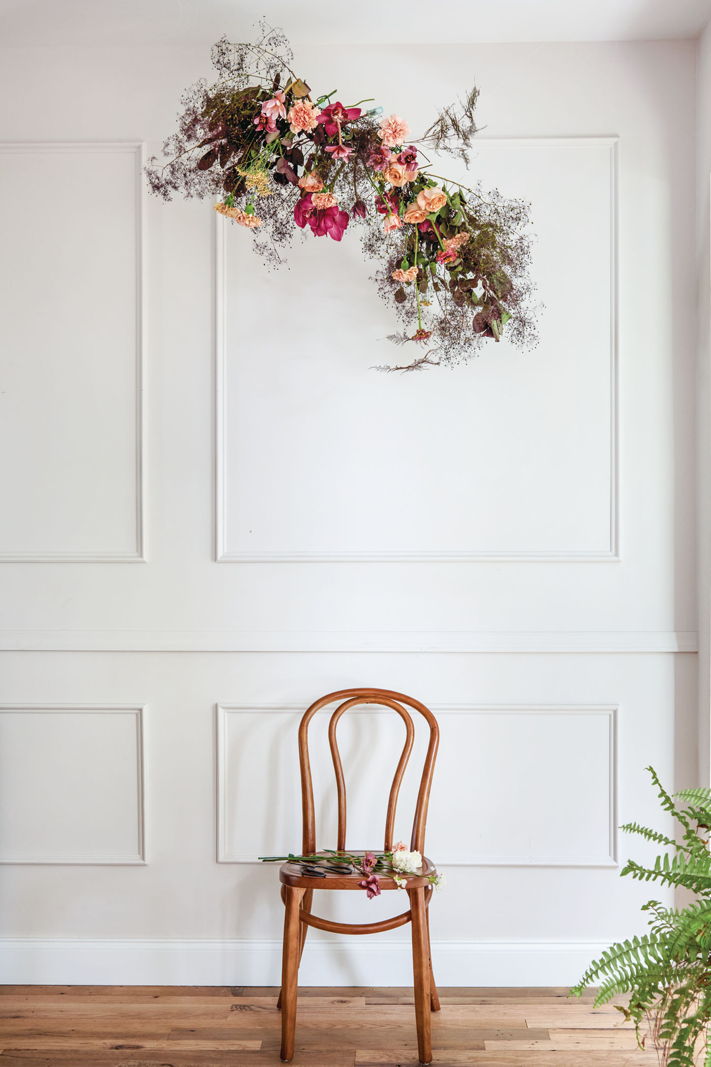 In a sunlight-filled, elegant white room, a suspended floral arrangement by Monica Delgado hangs above a simple wooden chair