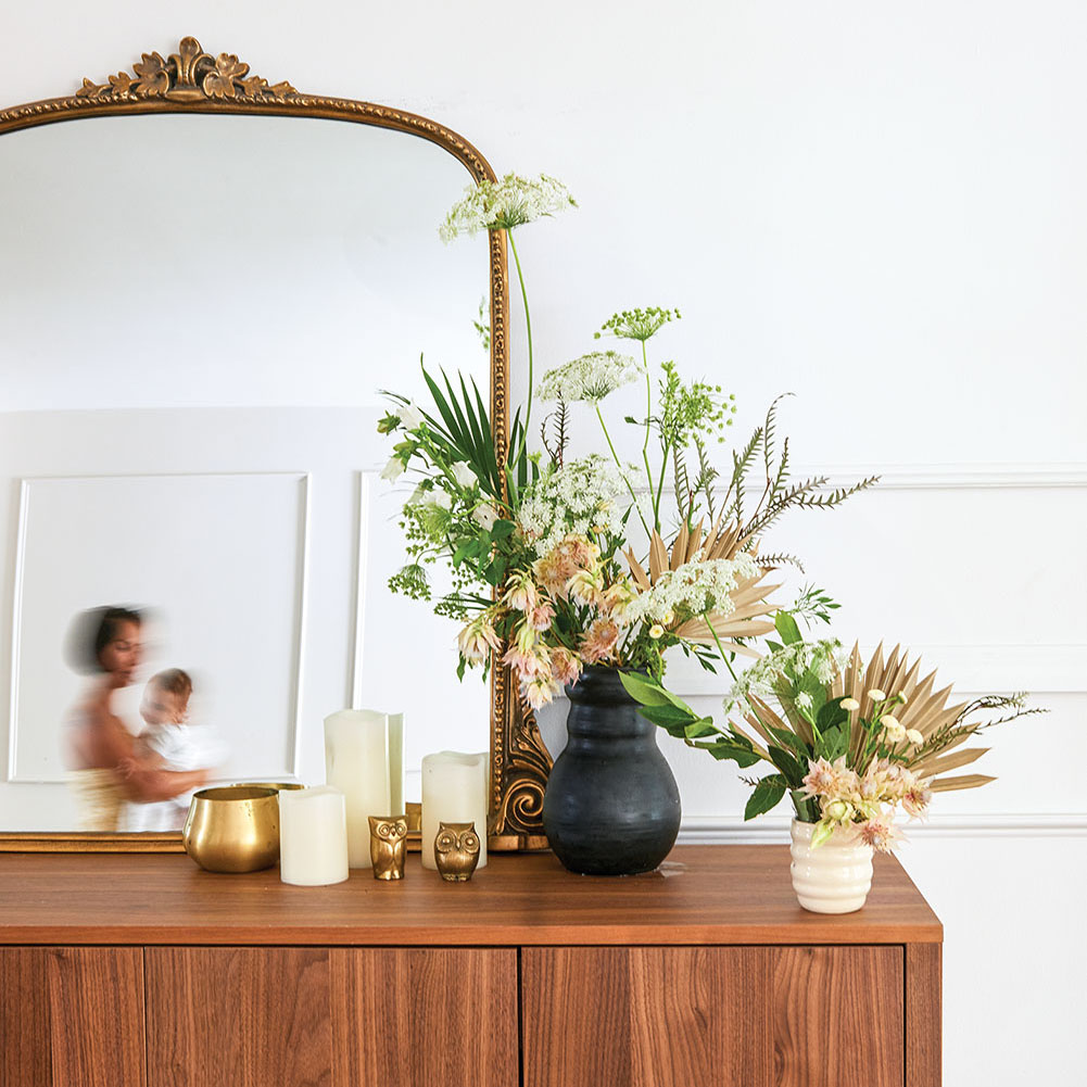 Two floral arrangements (one in a small white modern vase and the other in a larger, dark modern vase) sit to the right side on top of a mirror-topped wooden dresser. The mirror’s reflection shows a blurred image of Monica Delgado walking by holding her young child