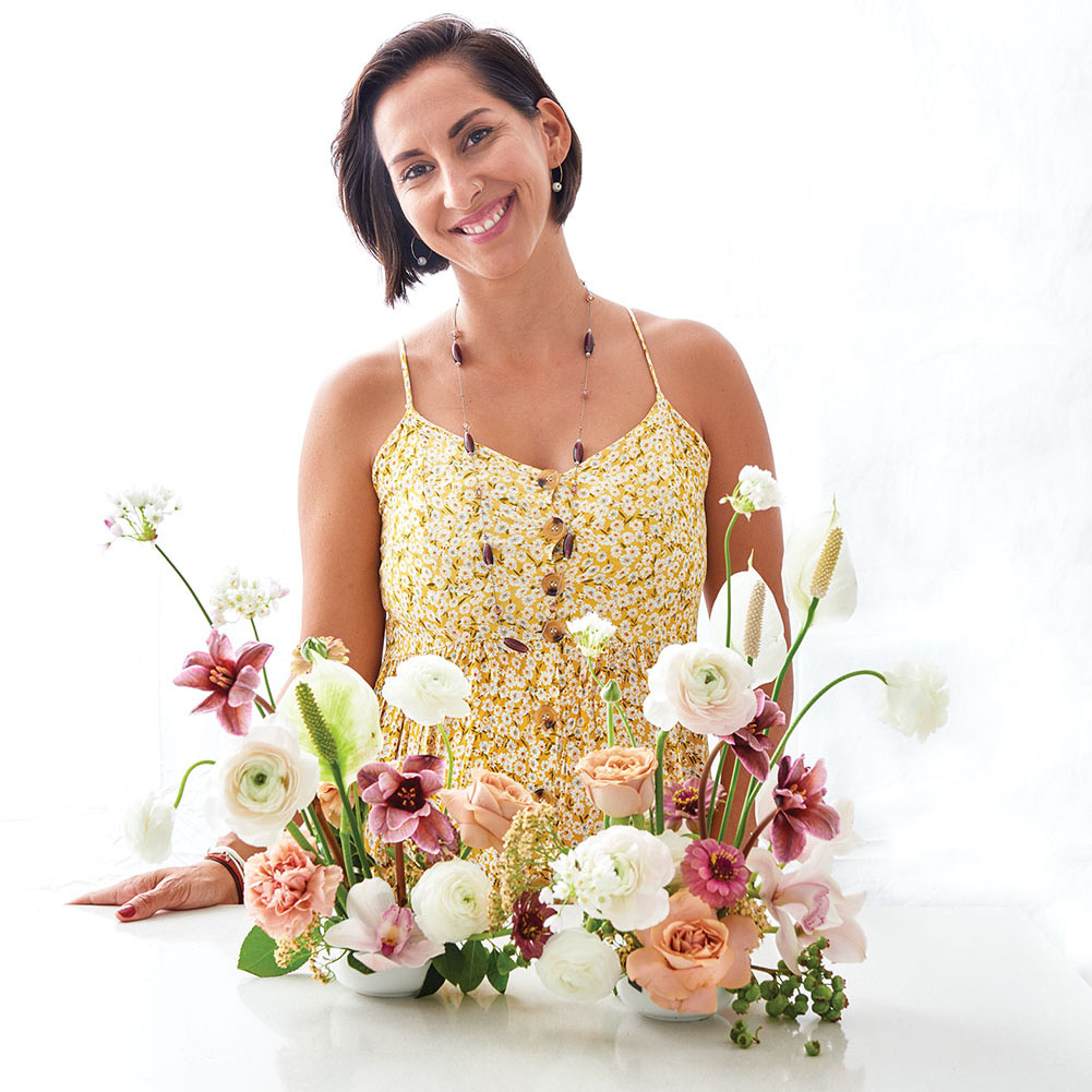 Portrait of floral designer Monica Delgado, with bobbed dark hair, wearing a yellow floral sundress and a long beaded necklace, stands with her ikebana arrangement
