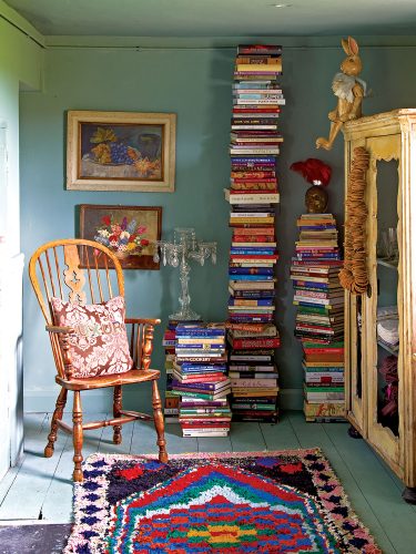 English country house interiors: Soaring towers of books in a cozy turquoise room with framed oil paintings, an antique wooden chair, and a colorful rug on a plank floor painted pale turquoise