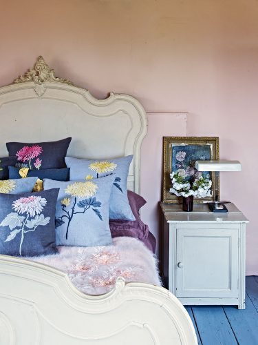 English country house interiors: pale pink bedroom with an ornate bed frame painted soft white, embroidered floral throw pillows, plank floors painted blue, and a bedside cabinet painted white and topped with a vase of flowers, an antique framed botanical oil painting, and a library-style reading lamp