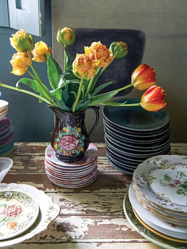 Yellow tulips in a vintage jug bearing a floral motif and stacks of Nikki Tibbles's vintage dinnerware on a rustic wooden tabletop with distressed white paint