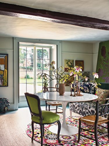 Nikki Tibbles's light-filled dining room features gracious French doors, an exposed ceiling beam, art-filled walls, layered rugs, and charming mismatched seating around the modern white circular table at the center