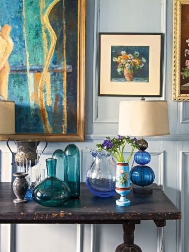 English country house interiors: a vignette of vintage glass and pewter vases on worn, dark wood table, with plentiful art, both modern and traditional, hanging on the pale gray walls behind