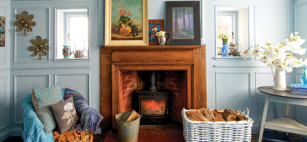In Nikki Tibbles' 1760 English country home, a fireplace with a warm-colored natural wood surround and mantle contrasts with pale blue-gray room (painted Farrow & Ball ‘Parma Gray’ wall). She has decorated the room with two solid bright blue plush rugs in contrasting textures, framed paintings, vintage vases, and a chair piled with throw pillows and cozy throws in cool colors. On a table to the right side of the room, large white enamel pitcher holds cut branches covered in white fluffy flowers. A large white basket holds chopped wood beside the fireplace.