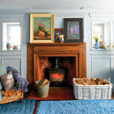 In Nikki Tibbles' 1760 English country home, a fireplace with a warm-colored natural wood surround and mantle contrasts with pale blue-gray room (painted Farrow & Ball ‘Parma Gray’ wall). She has decorated the room with two solid bright blue plush rugs in contrasting textures, framed paintings, vintage vases, and a chair piled with throw pillows and cozy throws in cool colors. On a table to the right side of the room, large white enamel pitcher holds cut branches covered in white fluffy flowers. A large white basket holds chopped wood beside the fireplace.