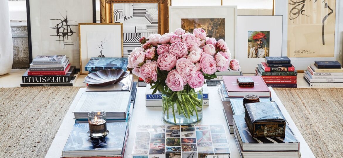 William McLure art and interiors: A large white square coffee table, with stacks of coffee table books on art and design and a vase of lush pink blooms at the center. Framed art is propped against the wall in the background