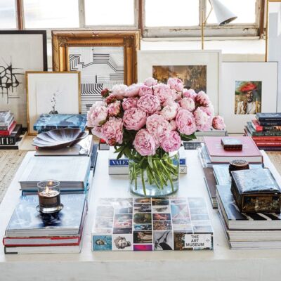 William McLure art and interiors: A large white square coffee table, with stacks of coffee table books on art and design and a vase of lush pink blooms at the center. Framed art is propped against the wall in the background