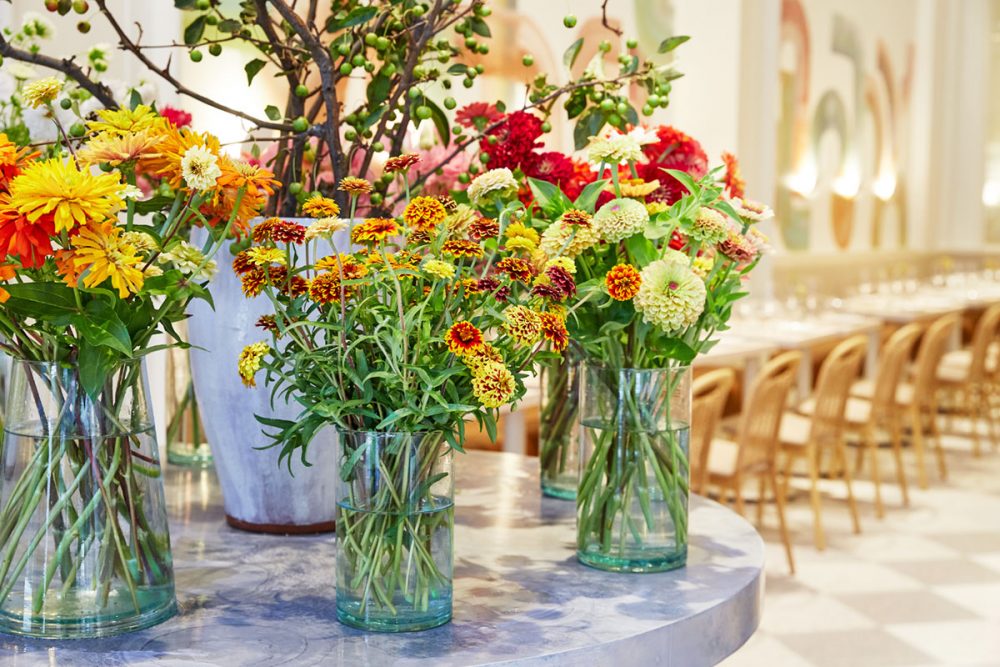 Close up view of vases of colorful zinnias standing on a round table at the entrance of Il Florista in New York City. In the background, you see a long row of tables and chairs on a white-and-pale gray diamond-patterned floor.