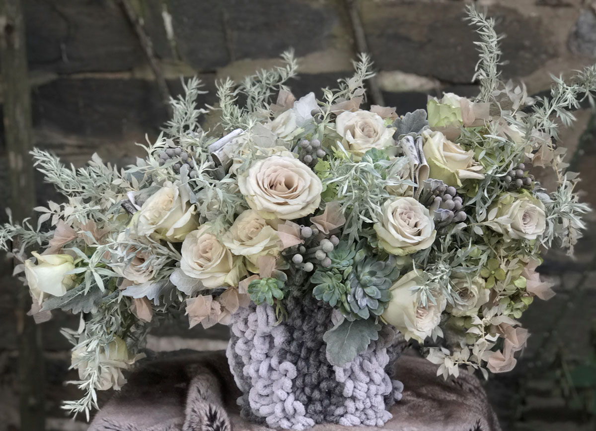 a floral arrangement featuring silvery foliage, muted roses, succulents, and a woven base in pale and charcoal grey yarn