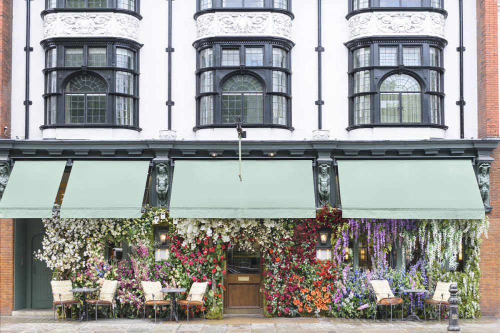 Beneath a green awning, the Ivy restaurant storefront is covered in colorful, lush blooms. Above, ornate windows overlook the London street scene 