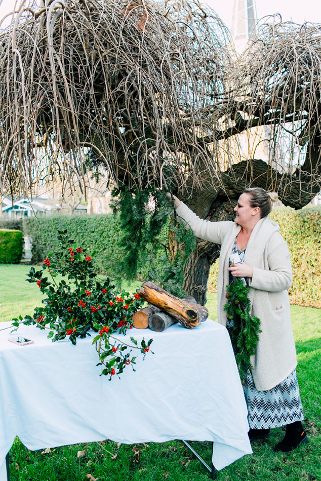 Floral designer Natalie Ransom hangs foraged evergreen foliage on a bare tree. Underneath, a table is decorated with holly branches. 