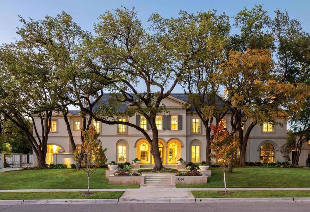 Warm yellow lights fill a grand beige mansion from a street view.