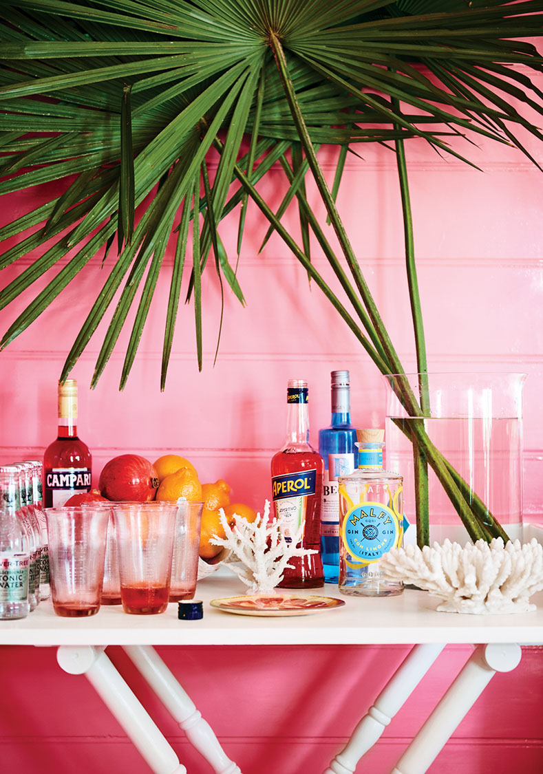 an at-home bar, with a vase of palm fronds, against a bright pink wall,
