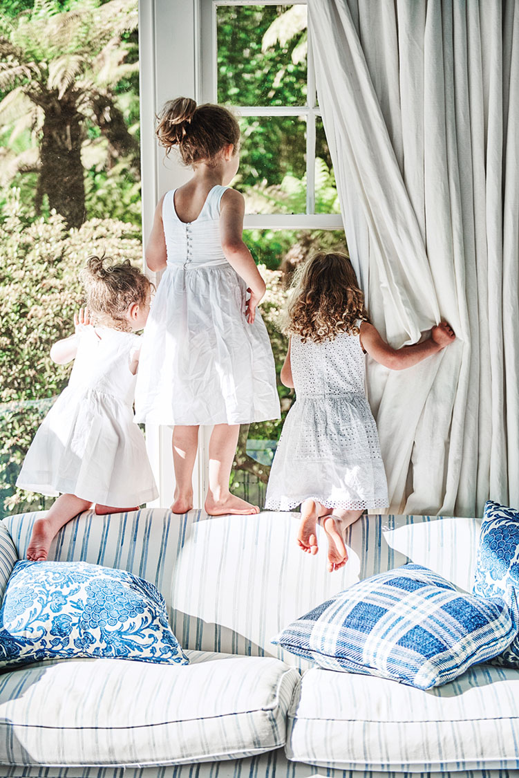 3 small children stand and kneel on a sofa back, peering through the window into the gardens