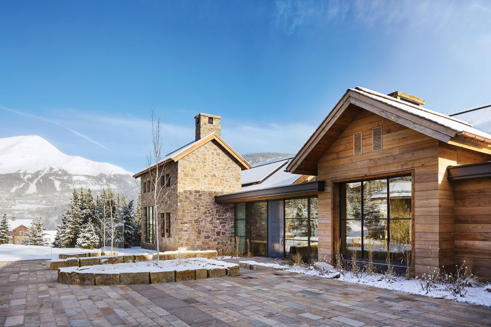 A mix of brick and wood make up the outside of a house sitting in snow-covered mountains.