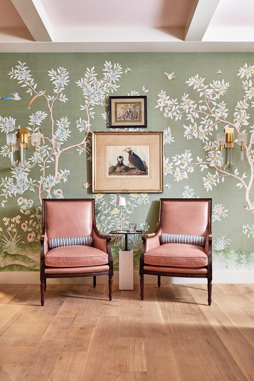 Pair of chairs in a shade of pink sit against green scenic wallpaper with pair of brass sconces in secret speakeasy designed by Corey Damen Jenkins