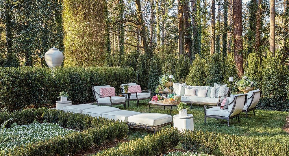 Sitting area in backyard between parterre garden and holly hedge. Sofa flanked by outdoor lamps and planters, four chairs, and four ottomans surround a round cocktail table set with flowers, fruit, and bottled sparkling water.