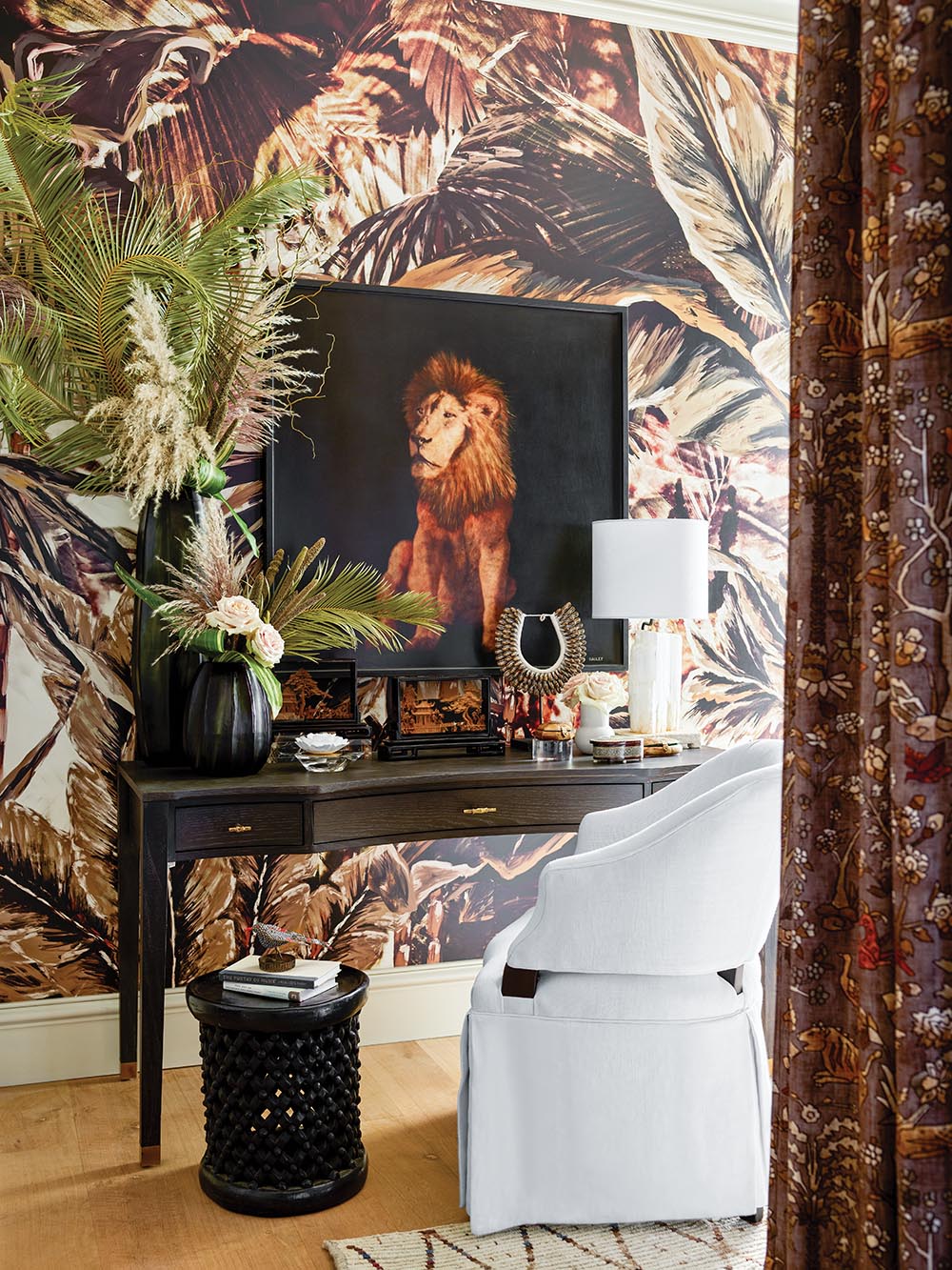 Desk with painting of lioness hanging atop jungle-print wallpaper. Tropical-inspired floral arrangements on desk.