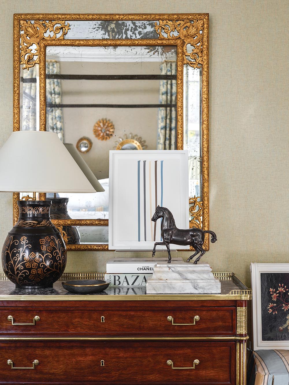18th-century chest topped with a 19th-century bronze horse, books, a lamp, and a nail polish painting by Scott Ingram. Gold-framed mirror.