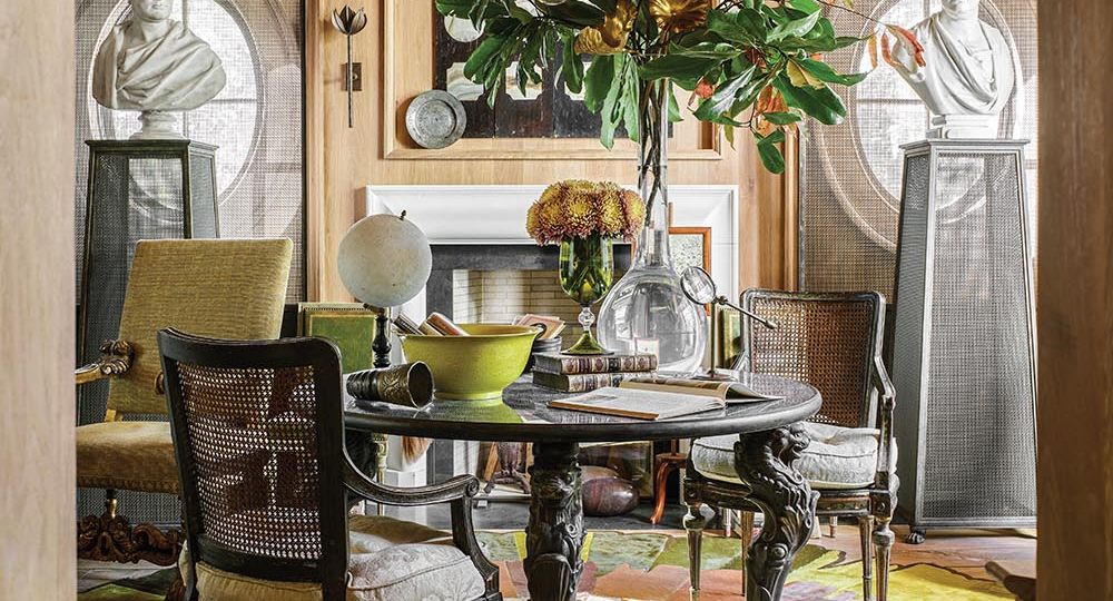 Table at center of home library designed by Barry Dixon. Dahlia rug on floor and vase of magnolia and other tree branches on table. Pair of busts on mesh pedestals in front of oval windows.